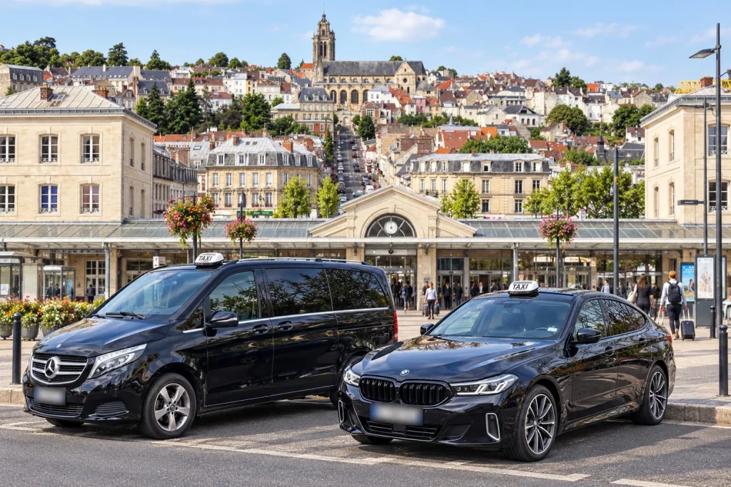 Un van Mercedes et une berline BMW taxis devant une gare, avec une ville en arrière-plan.